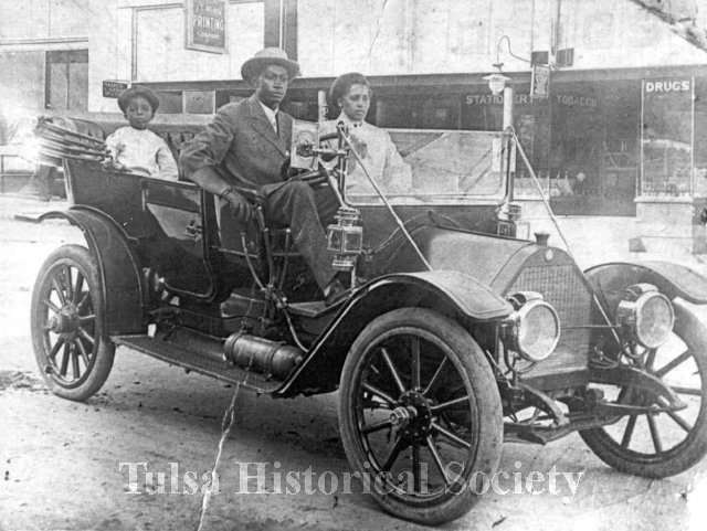 Reproduction, black & white photograph measuring 5" x 7" of John Wesley Williams and wife, Loula Cotten Williams, and their son, William Danforth Williams, sitting in their 1911 Chalmers touring car. A sign advertising A. L. Black Printing Company, located at 114 S. Boston Ave, Tulsa, OK, is visible in the background. John was an engineer for Thompson Ice Cream Company. Loula was a teacher in Fisher. The Williams family owned the Dreamland Theatre, which opened in 1914 at 129 North Greenwood Avenue, and was destroyed in the 1921 Tulsa Race Massacre.